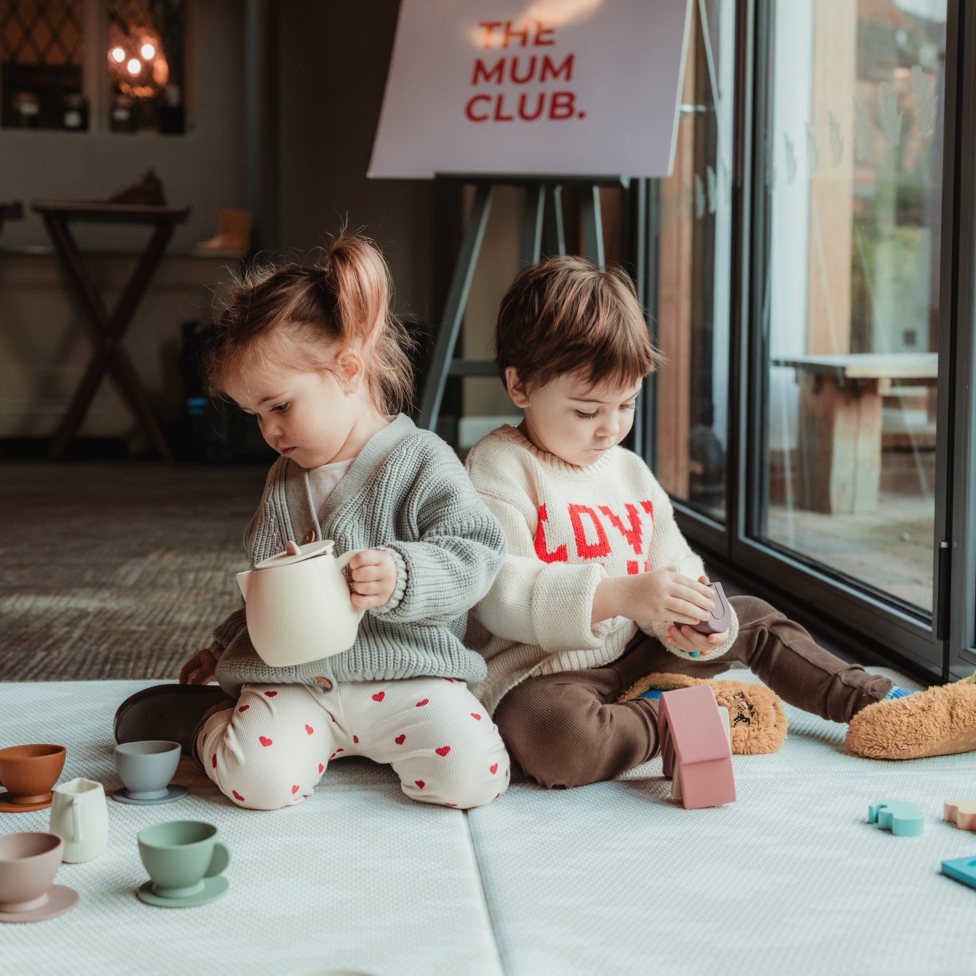 Two children playing with toys on a light-colored floor, with a sign reading 'The Mum Club' in the background.